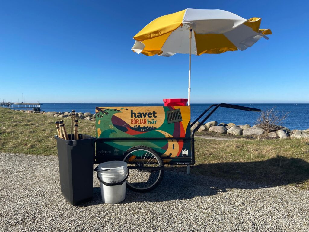 Vagn med strandstädarutrustning och med parasoll. På vagnen står det "Havet börjar här". I bakgrunden blå himmel och blått hav.