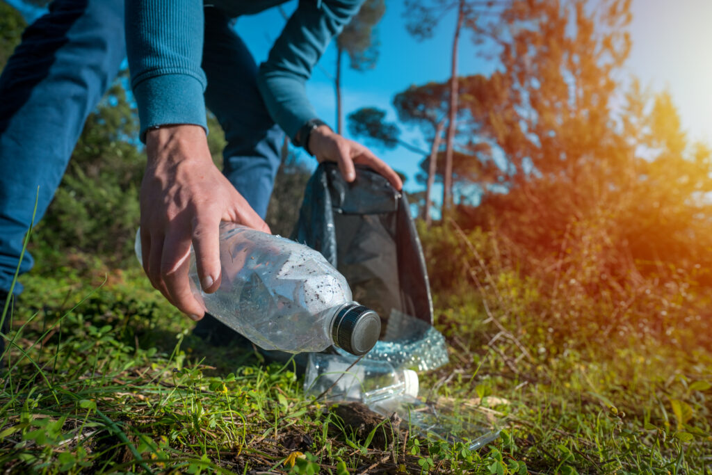 En person böjer sig ner och plockar upp en plastflaska och annat plastskräp i skogsterräng.